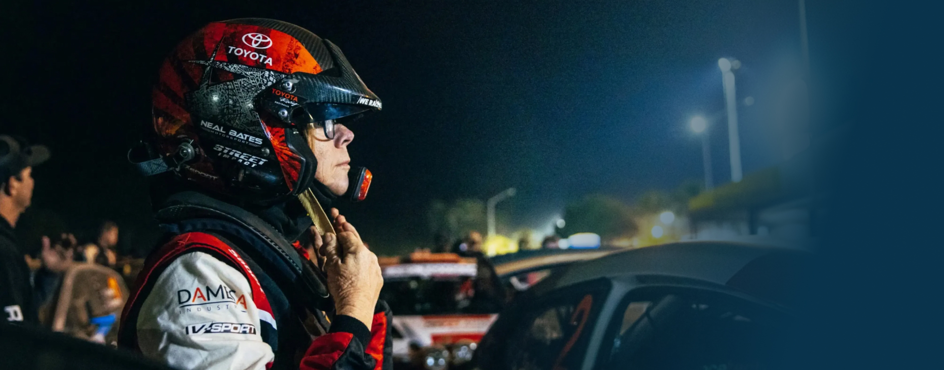 Coral Taylor standing next to her open racing vehicle, adjusting her helmet at a night rally. Other racers and observers dot the background, as stadium lighting illuminates the area.