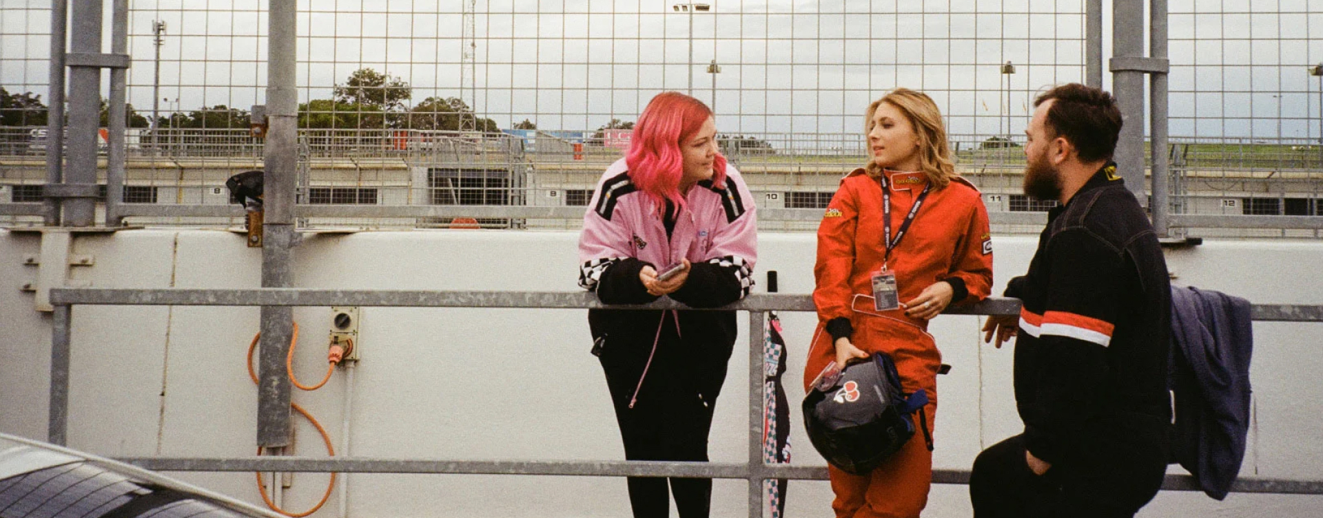 A G R C member chats with car racers and partners Jacinta and Kieran. Jacinta wears a bright orange racing suit, leaning casually against a steel handrail.