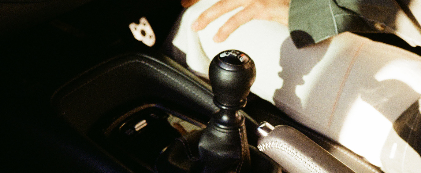 A close-up view of a 'G R' branded gearstick, showing the detailed white stitching along the centre console's black leather. A woman sits in the driver's seat, his hand and jacket sleeve visible, as the sun shines through the passenger side window.