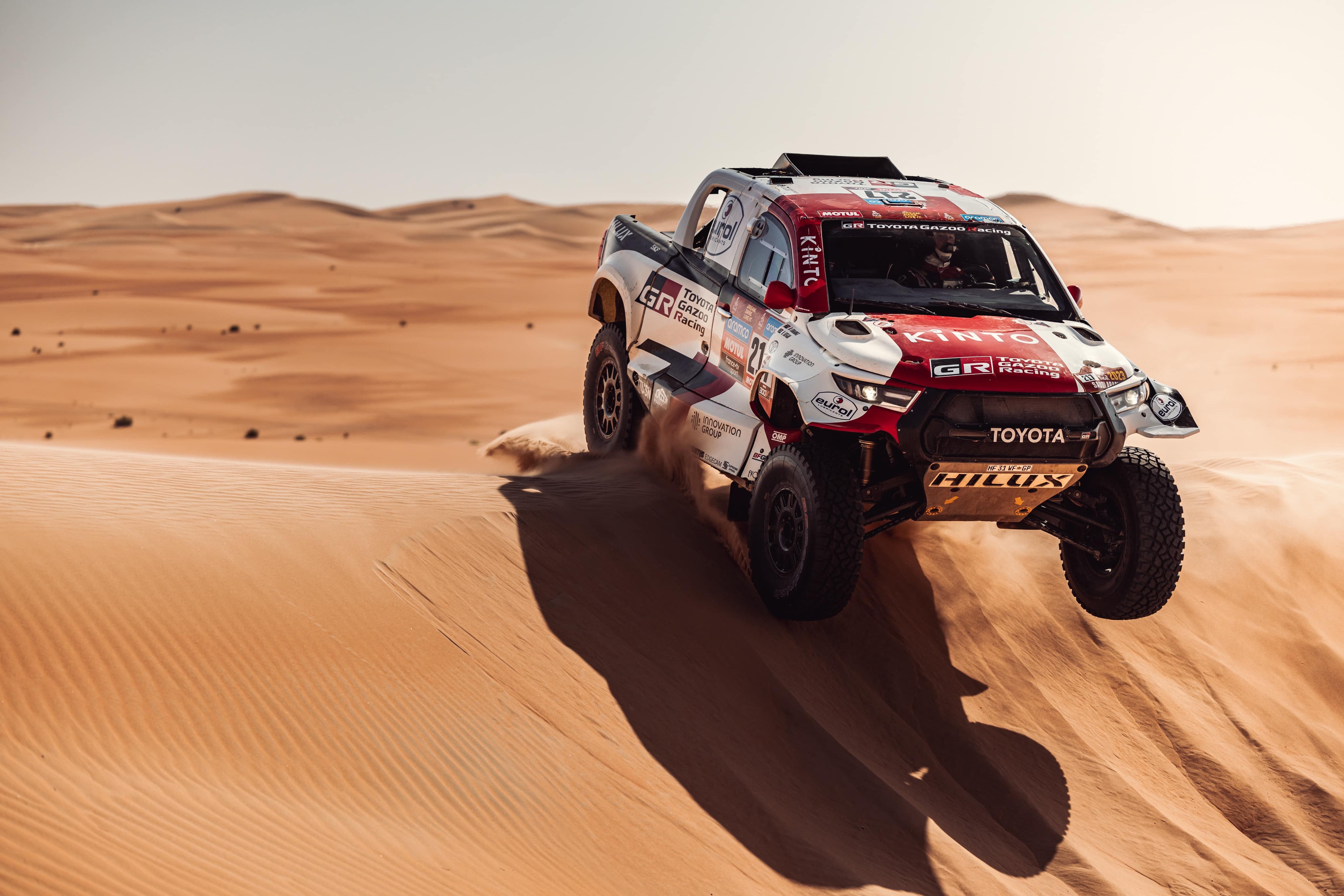 A Toyota G R Dakar HiLux covered entirely in red and white livery, its front wheels mid-air as it traverses over a large orange dune. The desert stretches out into the distance.