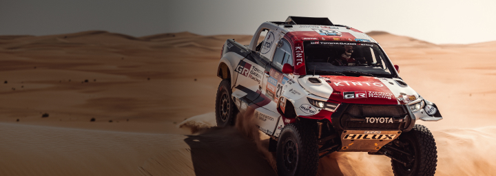 A Toyota G R Dakar HiLux covered entirely in red and white livery, its front wheels mid-air as it traverses over a large orange dune. The desert stretches out into the distance.