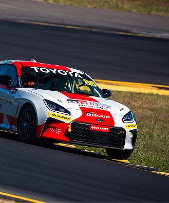 A Toyota GR86 in racing livery on a circuit, its right wheels just staying on the black bitumen as it comes around the bend.