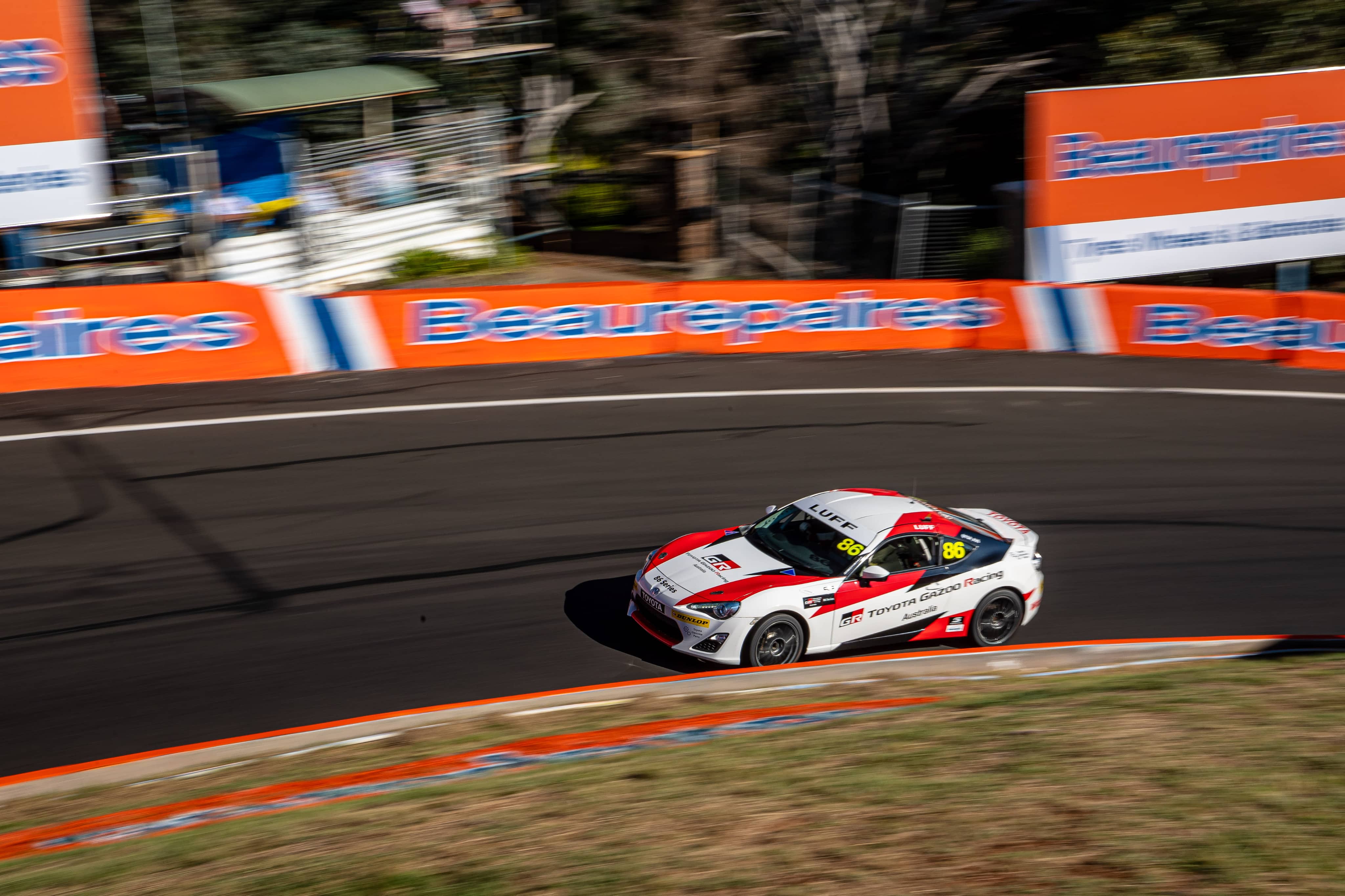 A Toyota G R 86 in red and white racing livery making the bend on a racetrack. The vehicle remains in focus while the surrounding environment is blurred in motion.
