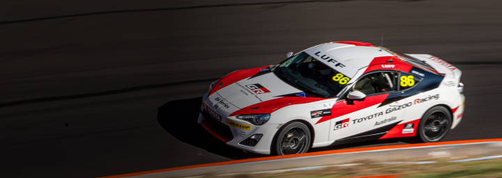 A Toyota G R 86 in red and white racing livery making the bend on a racetrack. The vehicle remains in focus while the surrounding environment is blurred in motion.