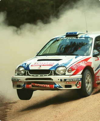 A Toyota G R Corolla decorated in red and blue racing livery featuring many sponsor brands. One wheel is in the air as it makes a sharp turn on a dirt rally track in the Australian bush.