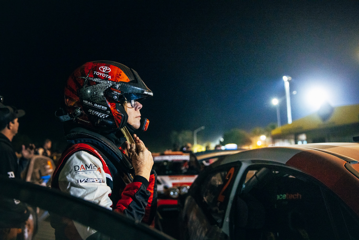 ARC champion Coral Taylor standing next to her open racing vehicle, fastening her helmet's chin strap at a nighttime racing event. Other racers, racecars and observers dot the background, as high-intensity lighting illuminates the area from a distance.