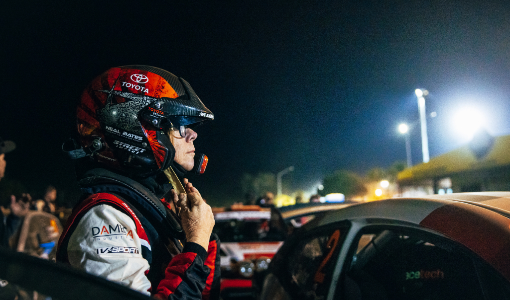 Coral Taylor standing next to her open racing vehicle, fastening her helmet's chin strap at a nighttime racing event. Other racers, race cars and observers dot the background, as high-intensity lighting illuminates the area. 