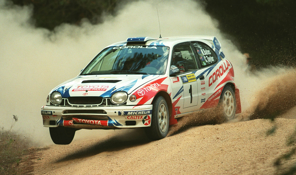 A Toyota Corolla WRC from 1996-1999 with Neal Bates and Coral Taylor at the helm. The front passenger side wheel is midair as it races around a rally track, kicking up gravel. The lower bumper and grille are damaged.