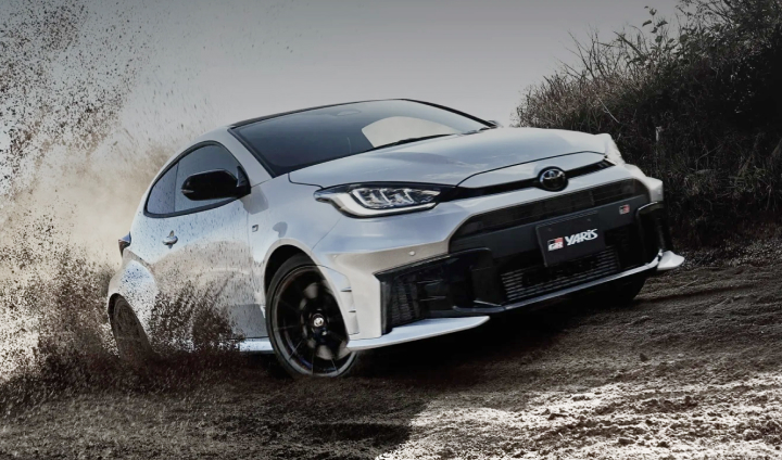 A dramatic angled shot of a Glacier White Toyota G R Yaris driving down a dirt track lined with long grass. The mud being kicked up by the tyres is arcing over the rear of the vehicle.