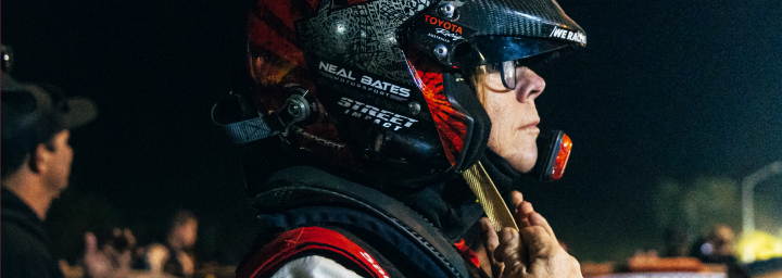 Coral Taylor standing next to her open racing vehicle, fastening her helmet's chin strap at a nighttime racing event. Other racers, race cars and observers dot the background, as high-intensity lighting illuminates the area. 