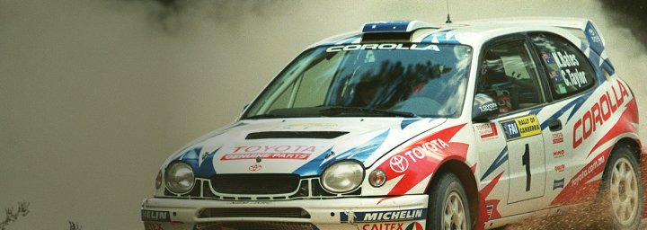 A Toyota Corolla WRC from 1996-1999 with Neal Bates and Coral Taylor at the helm. The front passenger side wheel is midair as it races around a rally track, kicking up gravel. The lower bumper and grille are damaged.