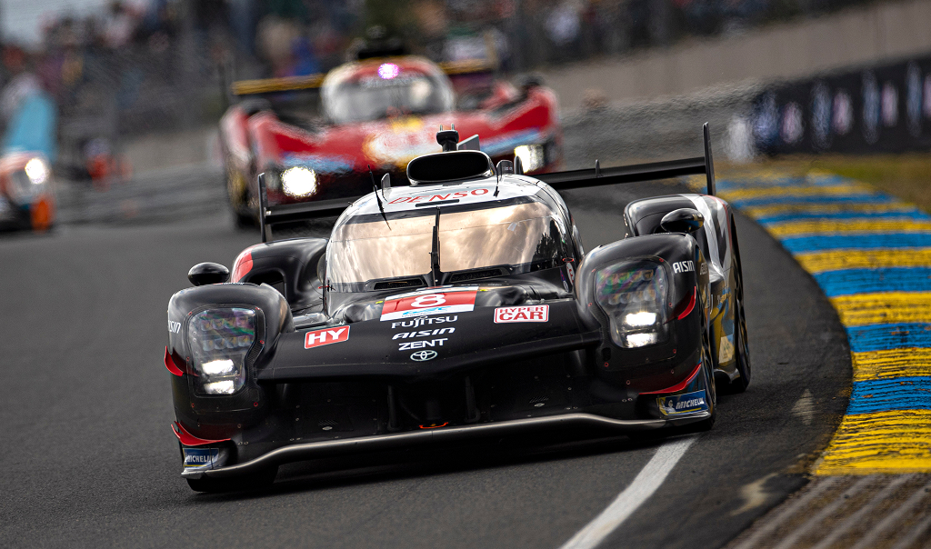 An angled view of a black Toyota G R 010 Hybrid Hypercar competing at Le Mans. The grandstand in the background is packed with people. 