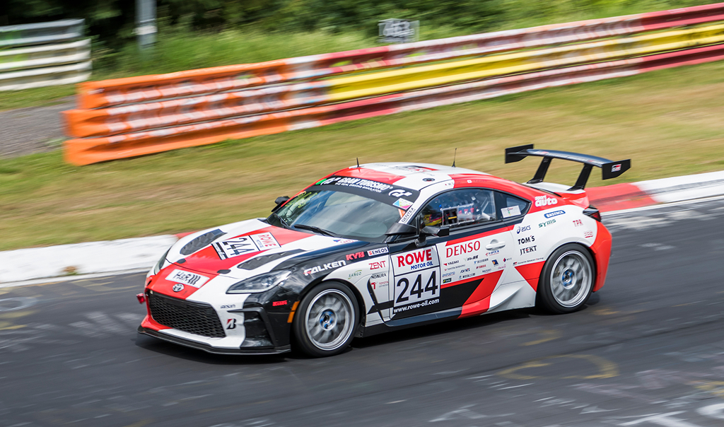 A White Liquid Toyota G R 86 with a large spoiler, in racing livery, speeding along a racetrack, its surroundings blurred in motion.