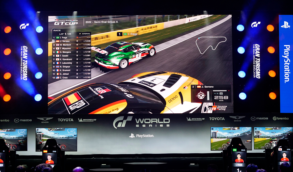A large screen showing a competitive e-sports race in the game Gran Turismo. Competitors sit below in front of monitors on gaming chairs.