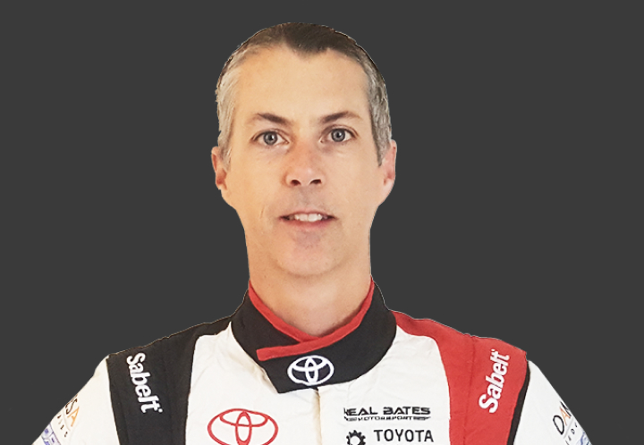 A headshot of professional T G R A co-driver Anthony McLoughlin in a white, black and red racing suit. Grey background.