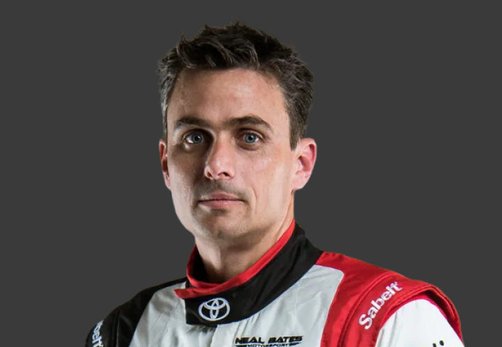A headshot of professional T G R A co-driver John McCarthy in a white, black and red racing suit. Grey background.