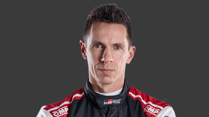 A headshot of world champion professional T G R co-driver Julien Ingrassia in a black racing suit with red and white detailing. Grey background.