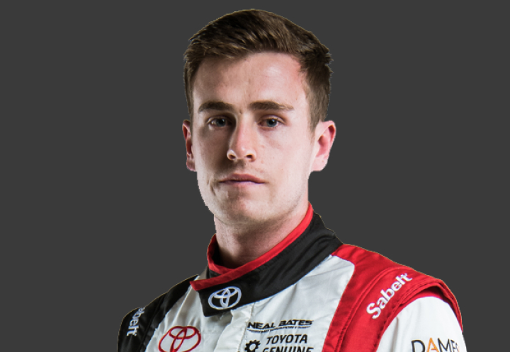 A headshot of professional T G R A driver Lewis Bates in a white, black and red racing suit. Grey background.