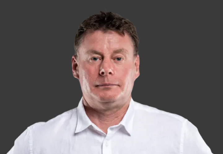 A headshot of T G R A Team Principal Neal Bates in a white suit shirt. Grey background.
