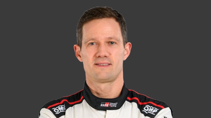 A headshot of world champion professional T G R driver Sébastien Ogier in a white racing suit with black and red shoulder details. Grey background.