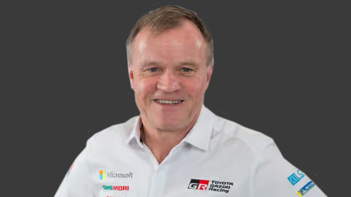 A headshot of world champion T G R Team Principal Tommi Mäkinen in a white T G R shirt. Grey background.