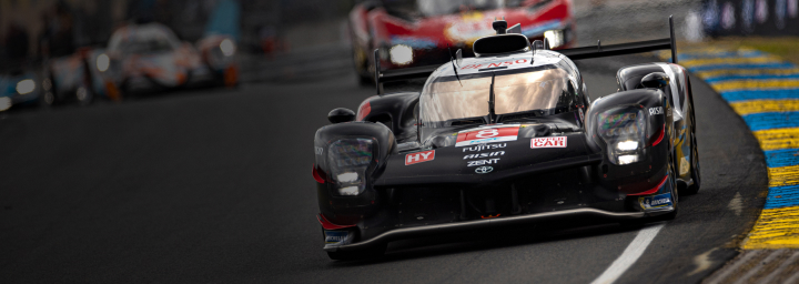 An angled view of a black Toyota G R 010 Hybrid Hypercar competing at Le Mans. The grandstand in the background is packed with people. 