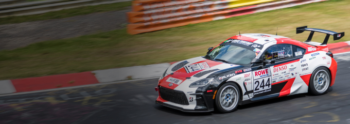 A White Liquid Toyota G R 86 with a large spoiler, in racing livery, speeding along a racetrack, its surroundings blurred in motion.