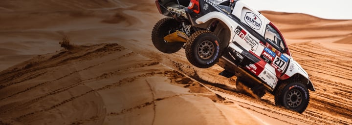 A Toyota Dakar HiLux in racing livery driving over the top of an orange dune. Its rear wheels are midair, nose turned toward the ground.