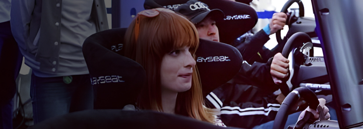 A large screen showing a competitive e-sports race in the game Gran Turismo. Competitors sit below in front of monitors on gaming chairs.