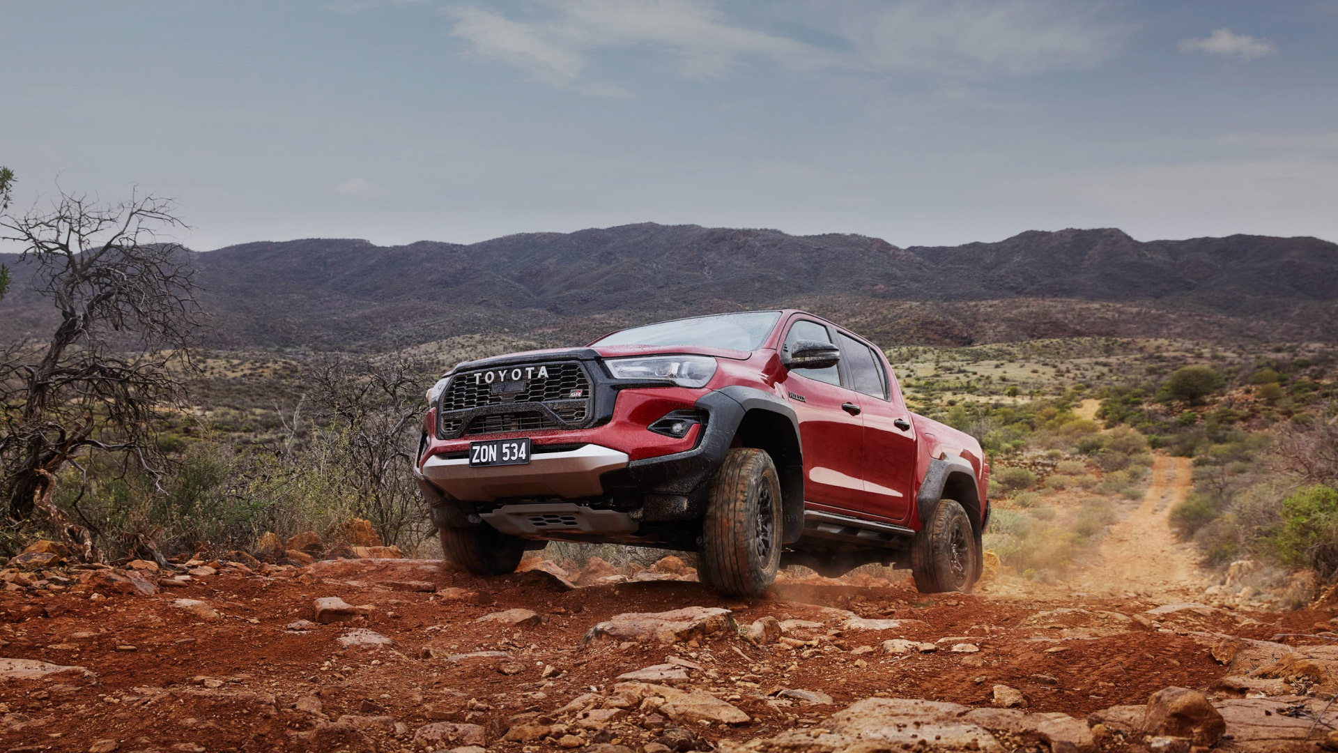 A red HiLux GR Sport climbs a steep off-road track in the mountains, kicking up red dust.