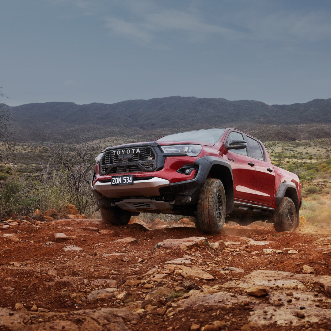 A red HiLux GR Sport climbs a steep off-road track in the mountains, kicking up red dust.
