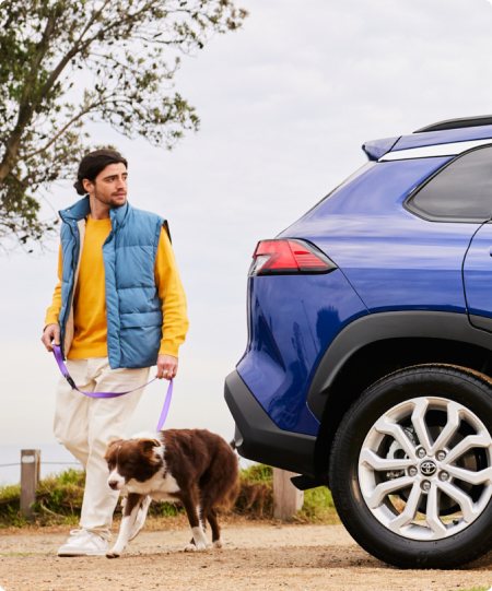 Young brunette man with a brown and white dog on a leash, walking away from a blue Toyota Corolla Cross.