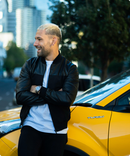 Young, smiling blonde man leans against a yellow GR Yaris.