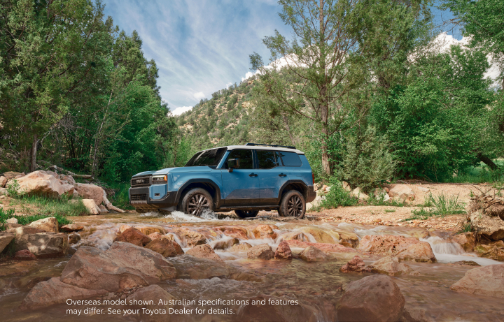 A blue LandCruiser Prado on a rocky river crossing surrounded by trees and mountains. 