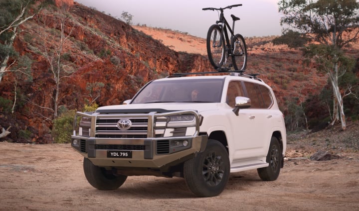 A glacier white coloured LandCruiser 300 Sahara with an alloy bull bar with/light bar and pro ride bicycle carrier on a dirt track.