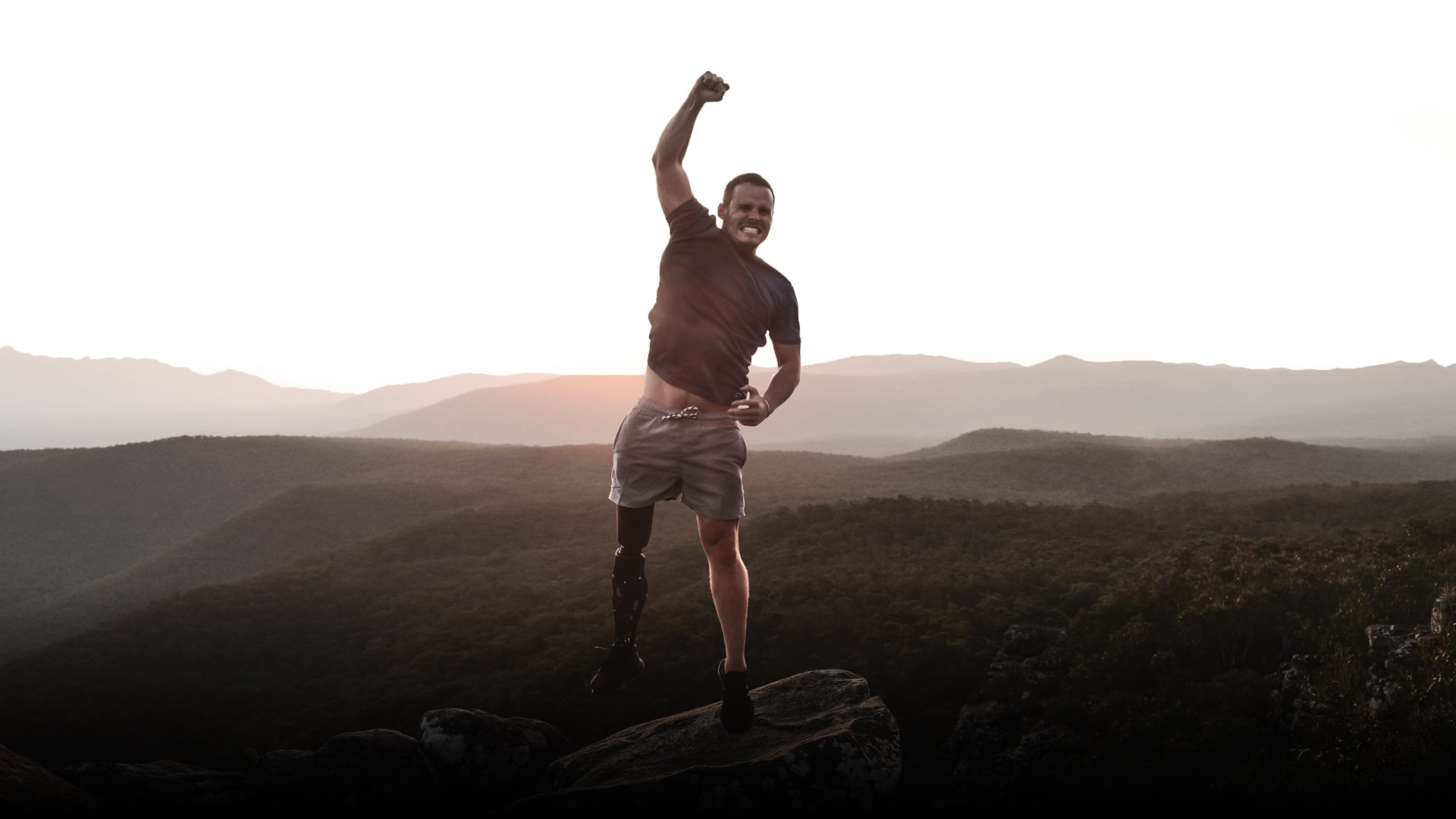 A tall, fit man with a bionic leg punches the air on a mountain at dusk.