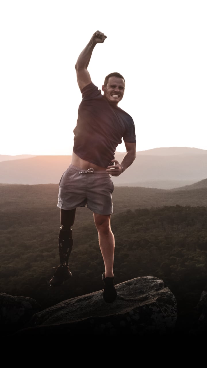 A tall, fit man with a bionic leg punches the air on a mountain at dusk.