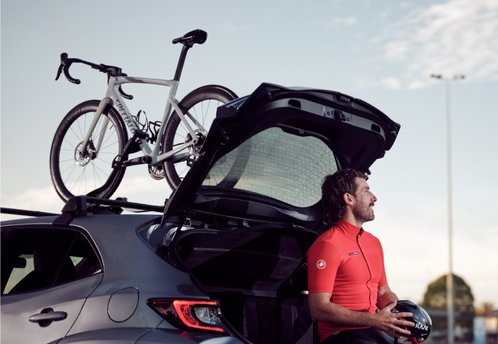 A man holds a bicycle helmet as he sits on the open boot of a grey car with a road bike on the roof rack.
