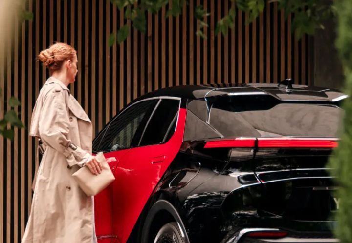 A woman is opening the door of a black and red Toyota C-HR parked next to a timber-slat fence.