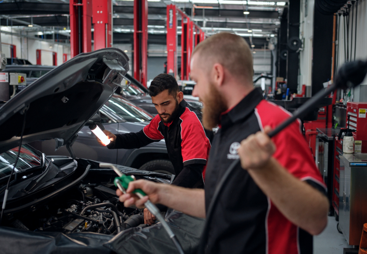 A man in a red and black Toyota uniform holds a light over an open engine while another man holds a nozzle in the foreground of a workshop.