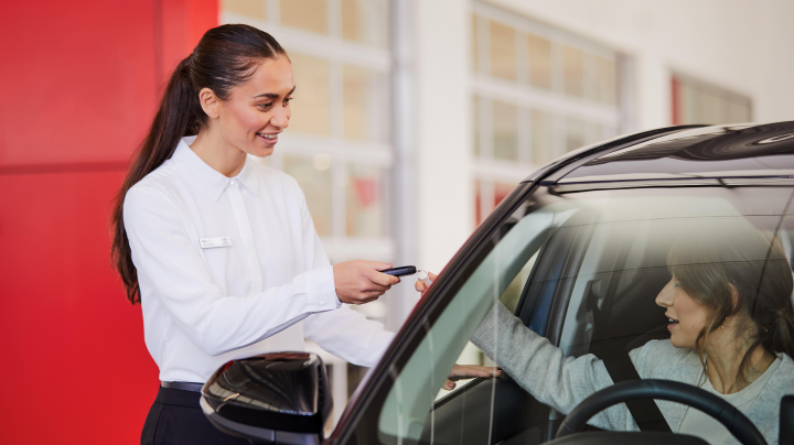 A woman smiles as she hands a car key to a woman sitting in the driver's seat of a black car inside a car showroom.