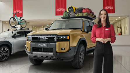 A woman in a red Toyota shirt stands next to a gold Toyota LandCruiser Prado with a loaded roof rack. Next to it is a grey car carrying a bicycle on its roof rack.