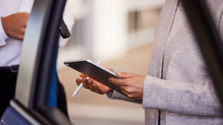 A person holds a tablet and a stylus standing next to a car door.