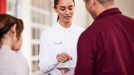 A woman speaks with a man and woman as she gestures toward a tablet.