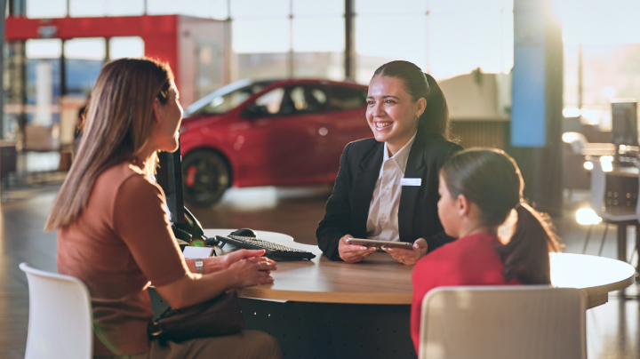 A woman in a blazer with a name badge sits at a round table smiling at a woman and a young girl inside a Toyota dealership.