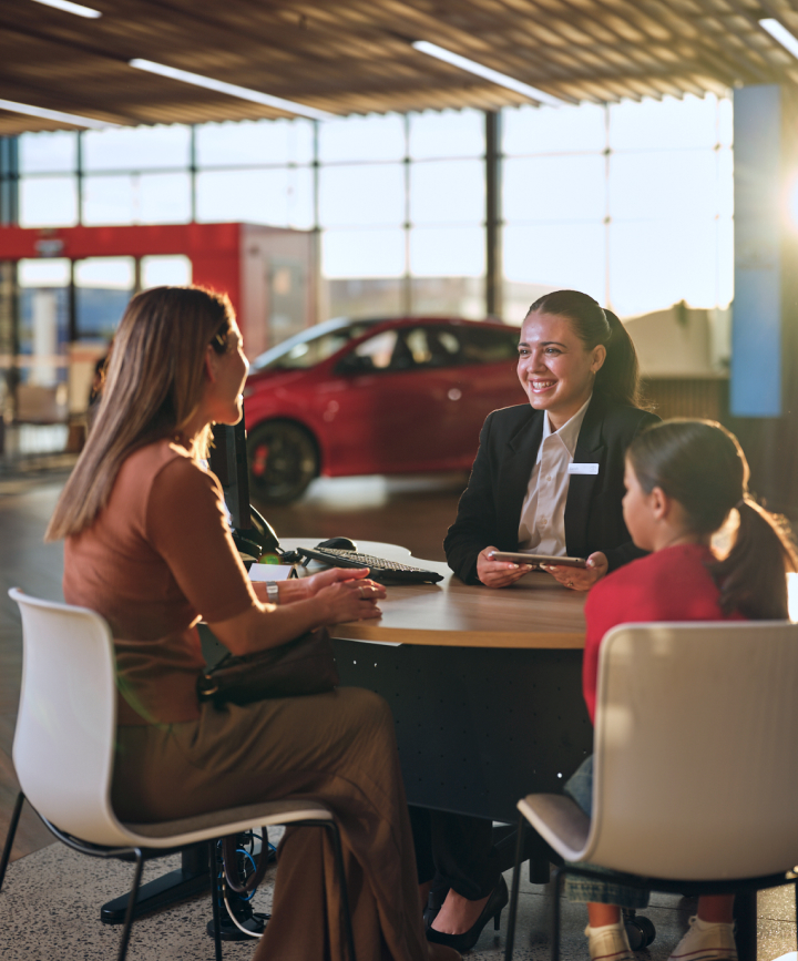 A woman in a blazer with a name badge sits at a round table smiling at a woman and a young girl inside a Toyota dealership.