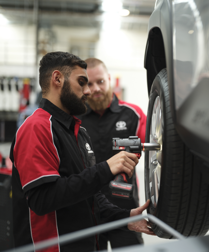A man in a red and black Toyota uniform uses a rattle gun to secure a wheel on a hoisted car. Another man looks on in the background.