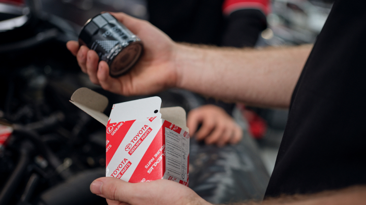 A close-up of a person holding a new black cylindrical filter in one hand and its open red-and-white 'Toyota Genuine Parts' box in the other.