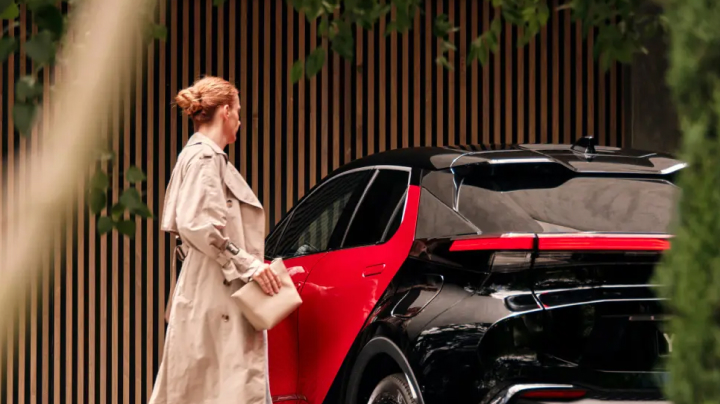 A woman is opening the door of a black and red Toyota C-HR parked next to a timber-slat fence.