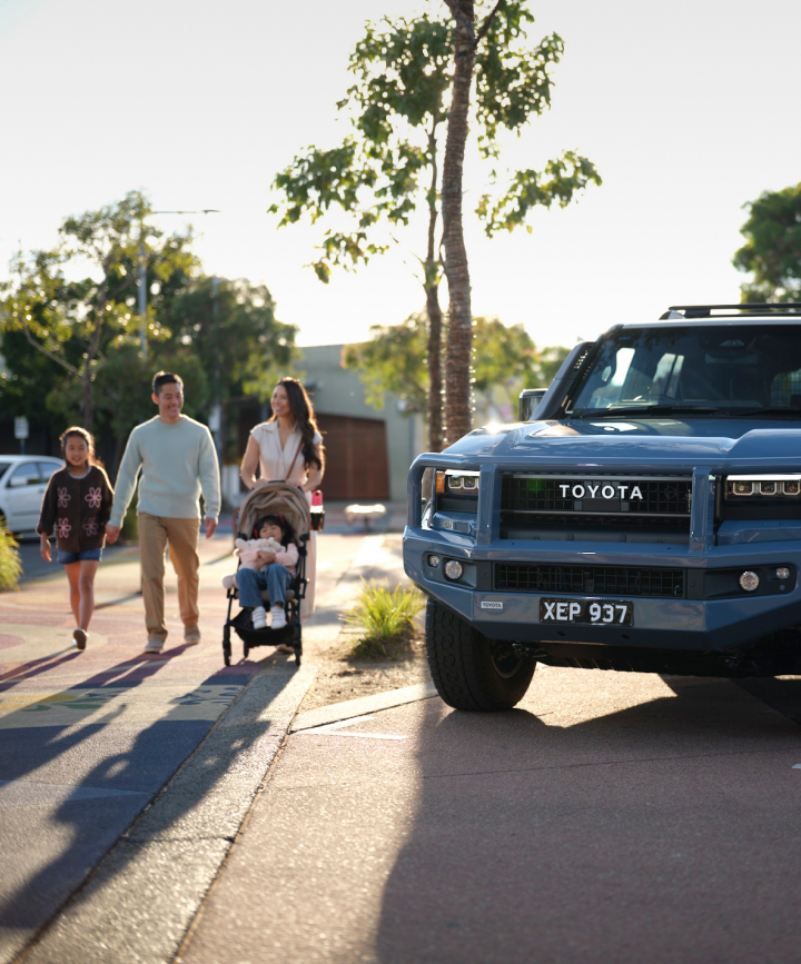 A blue Toyota Prado is parked on a suburban street. A man, a woman, a young girl, and a baby in a pram walk on the footpath in the background.